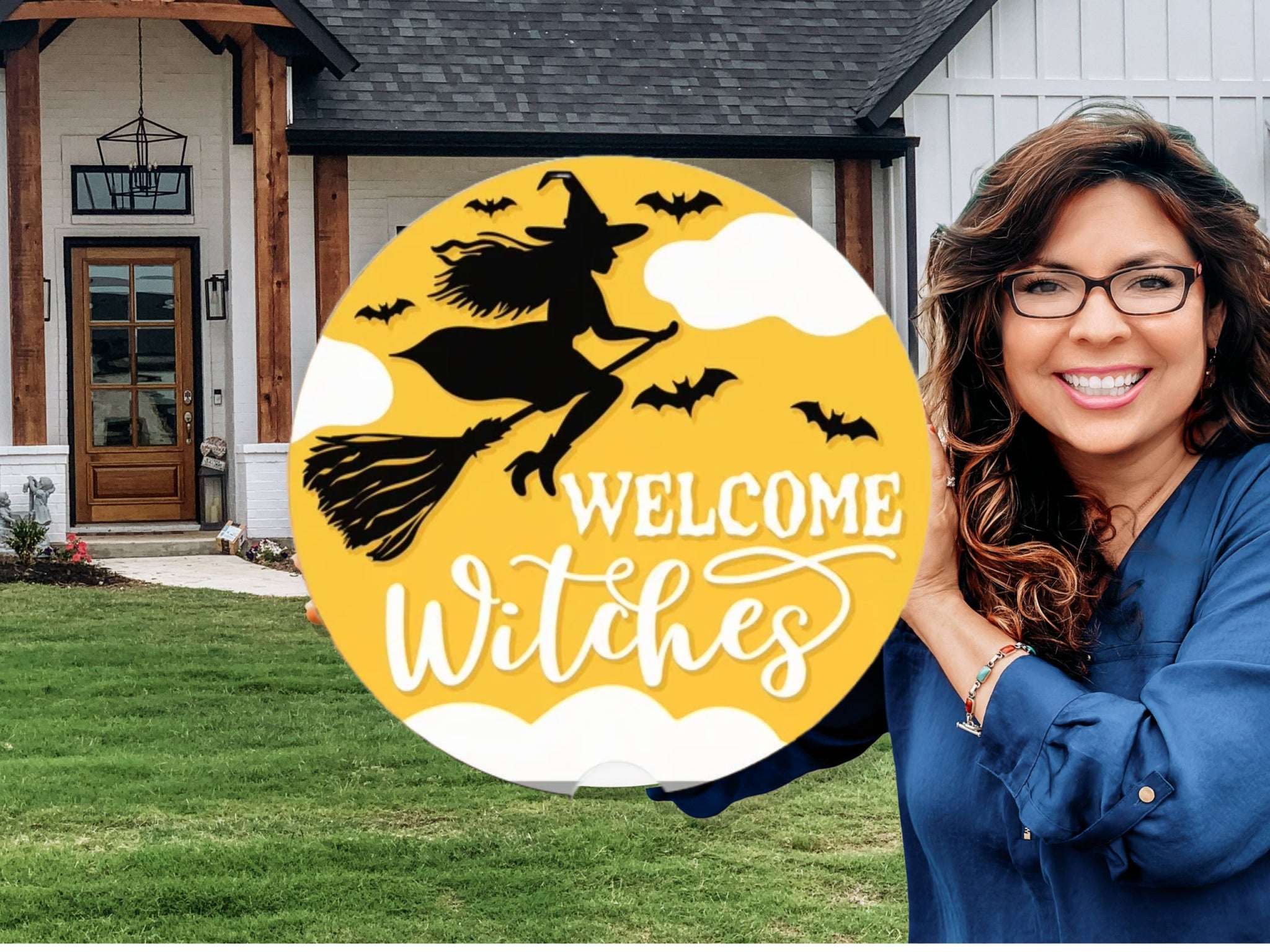 A smiling woman stands outside a house holding a round yellow sign with a silhouette of a witch on a broomstick, bats, clouds, and the words “Welcome Witches” in white script.