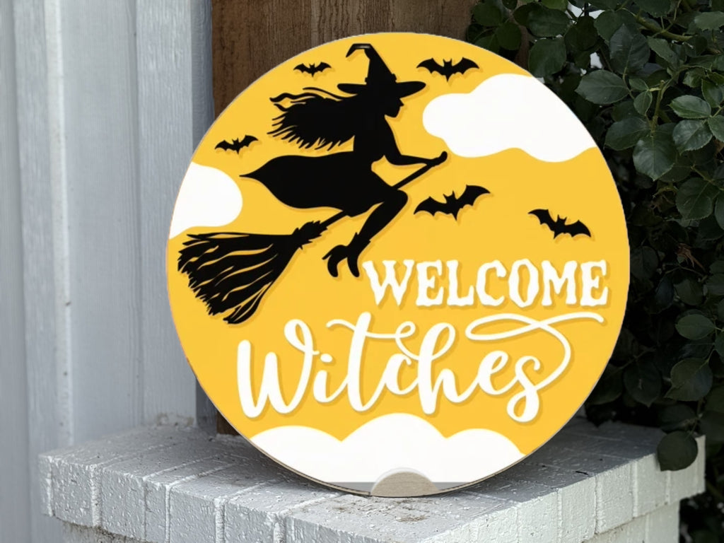A round yellow sign with a black silhouette of a witch on a broomstick, flying among bats and clouds, with the white text Welcome Witches. The sign is propped against a wall near some greenery.