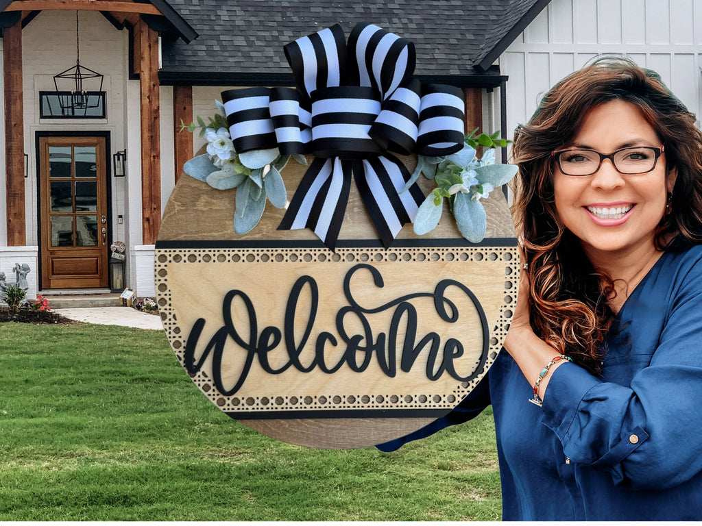 A smiling woman stands in front of a house, holding a round wooden sign with a striped bow, greenery, and the word “Welcome” written in cursive.
