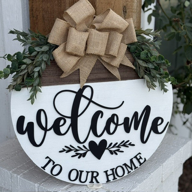 A round wooden sign with Welcome to Our Home in black script hangs on a wall. It features a heart with leafy accents. Atop the sign is a decorative burlap bow surrounded by green foliage, adding a rustic touch. The backdrop shows a partial view of a wooden wall and white brick.