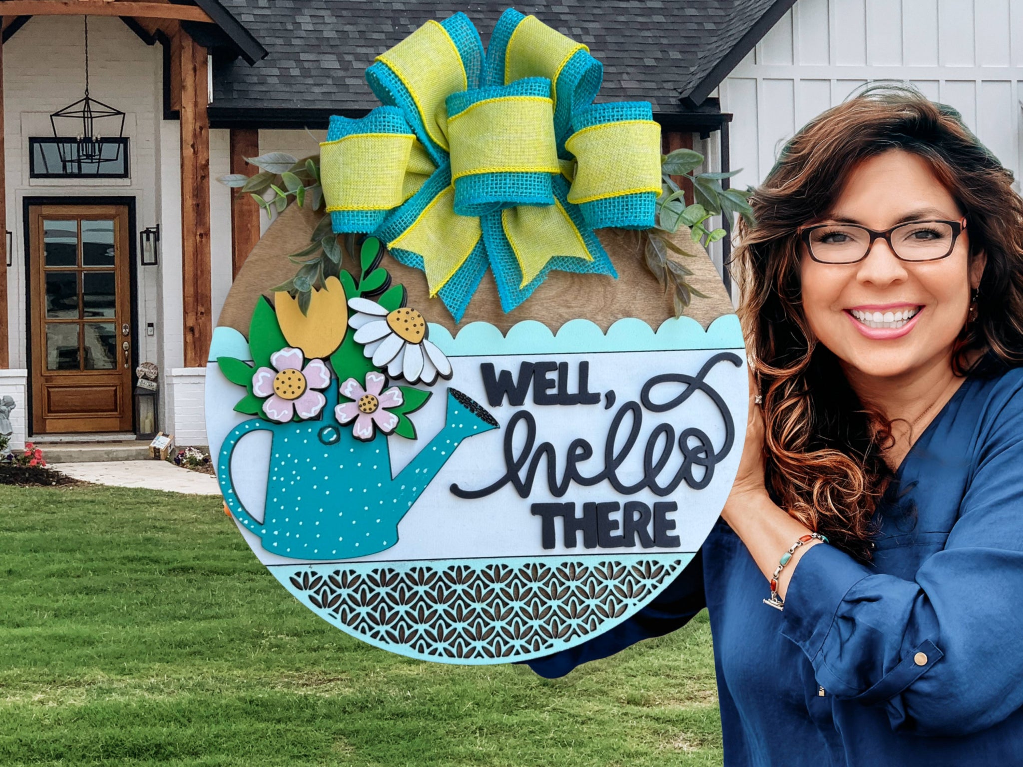 A smiling woman in glasses and a blue shirt holds a decorative round sign with a big bow, flowers, and a watering can, reading “Well, hello there,” standing outside a modern house.