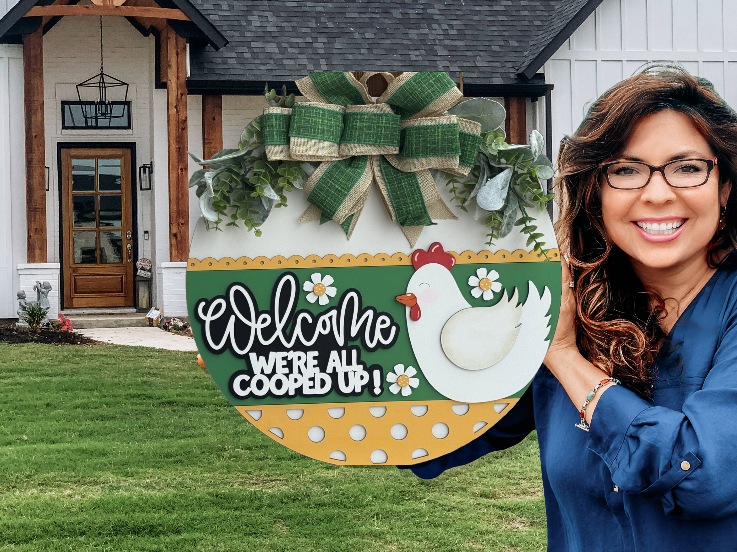 A smiling woman holds a round, decorative sign with a chicken, flowers, and the words Welcome, Were All Cooped Up! outside a modern farmhouse with a green lawn and wooden accents.
