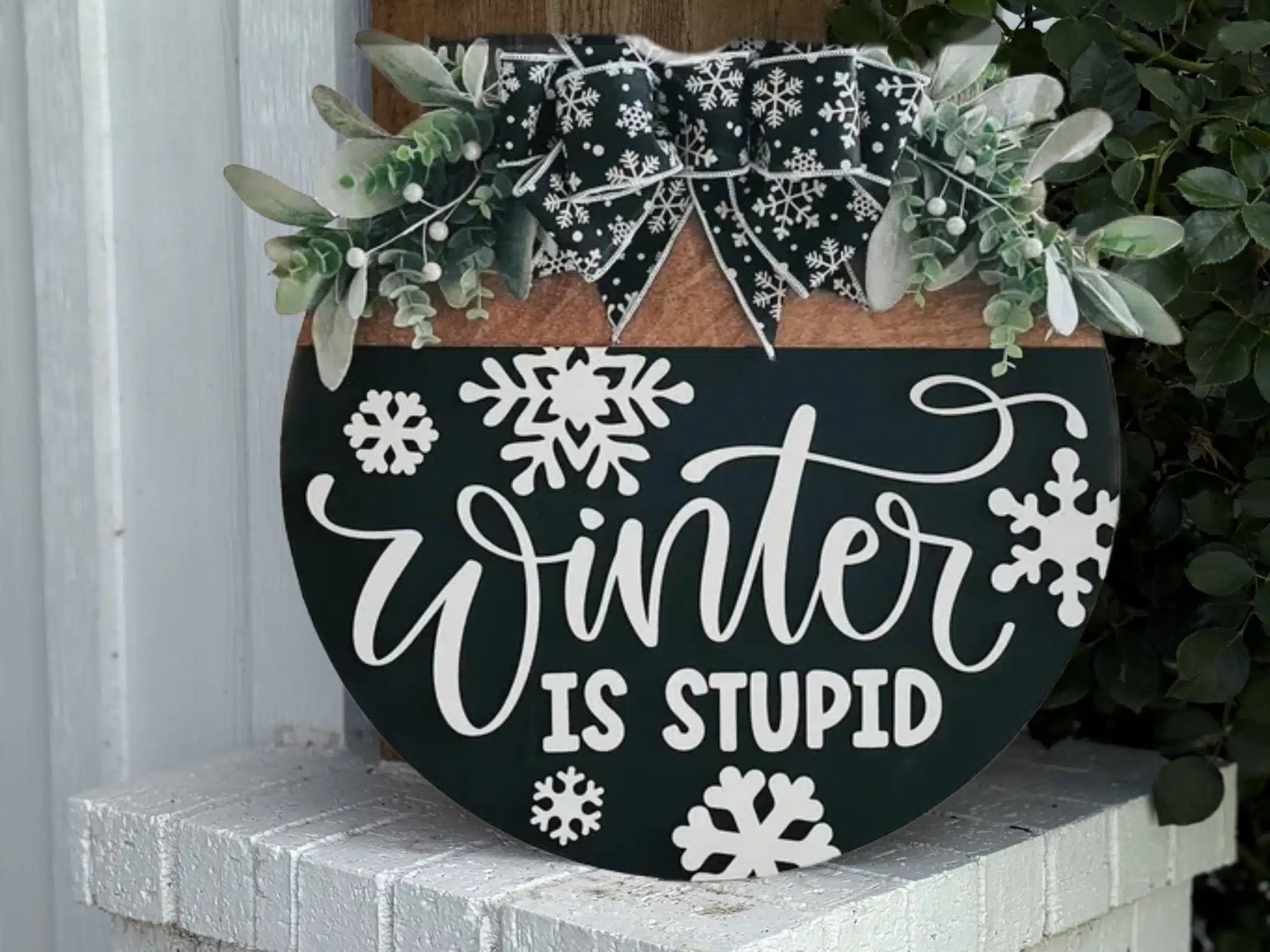 A decorative sign with white snowflakes and the words “Winter is Stupid” in cursive, topped with greenery and a black and white plaid bow, sits by a white brick wall and leafy plant.