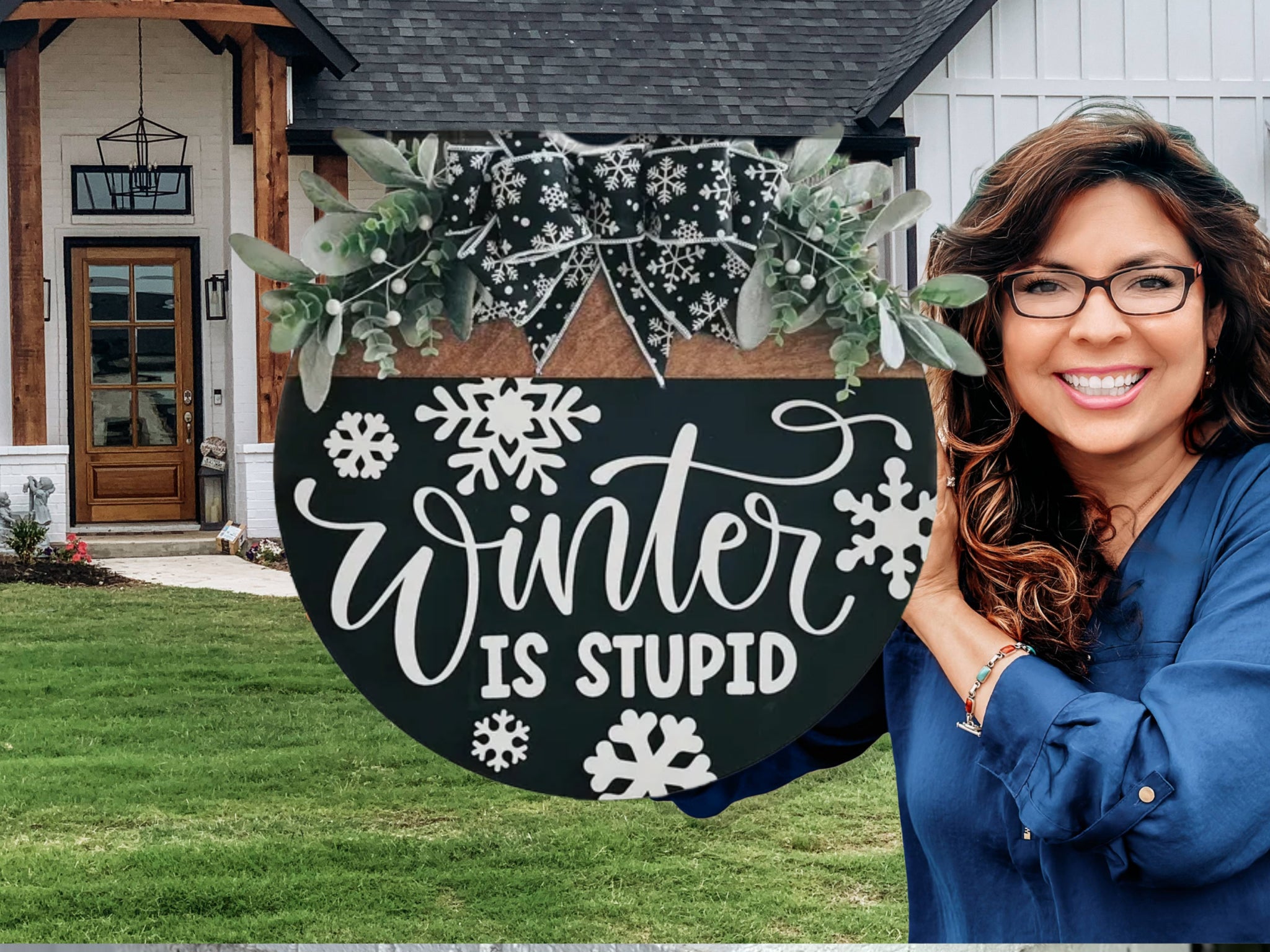 A smiling woman with wavy brown hair and glasses holds a round sign decorated with snowflakes, greenery, and a bow. The sign reads Winter is Stupid. She stands in front of a modern house with a lawn.