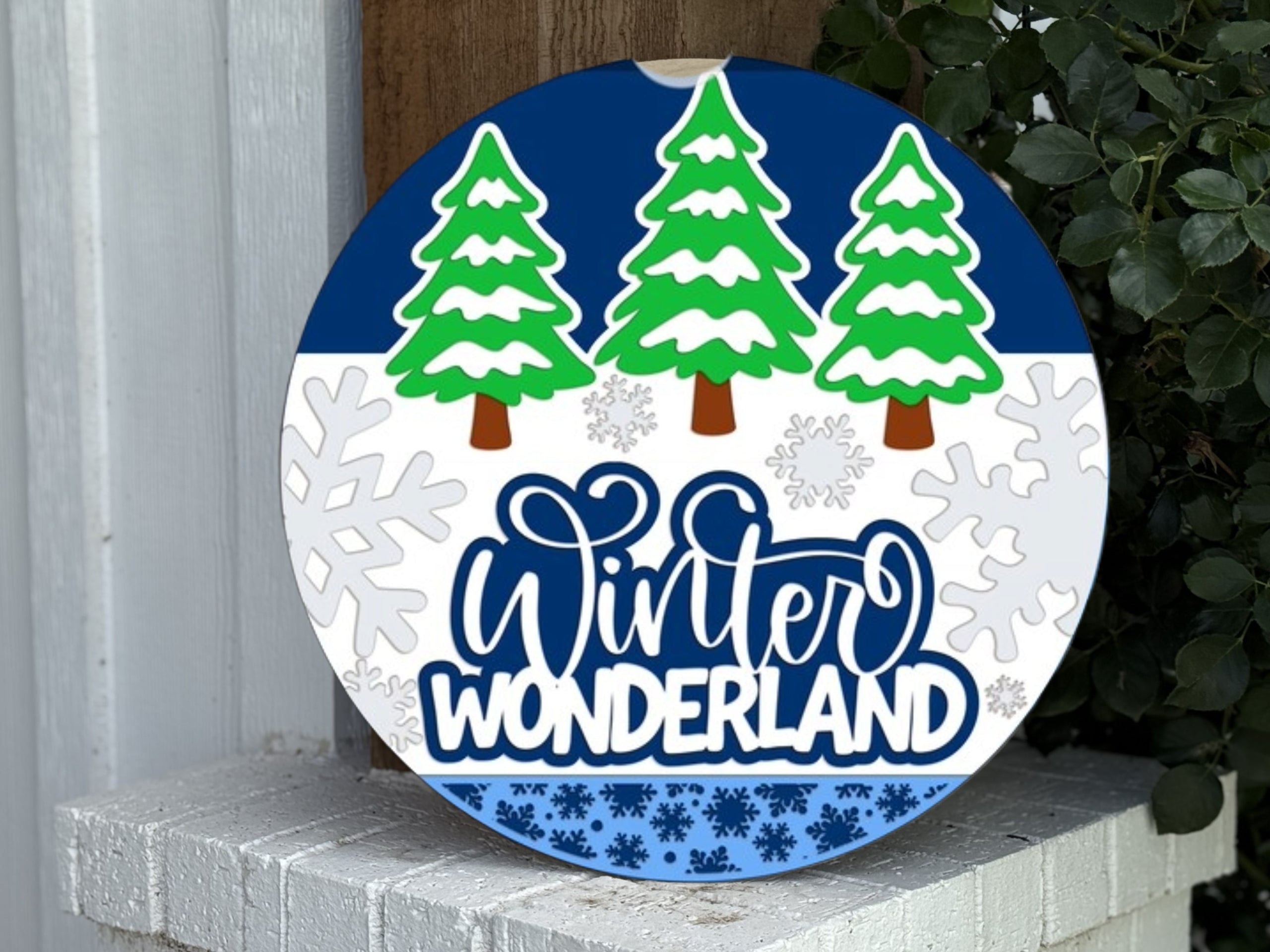 A round sign with the words Winter Wonderland, decorated with green pine trees and white snowflakes on a blue and white background, displayed outdoors near greenery and a white brick surface.