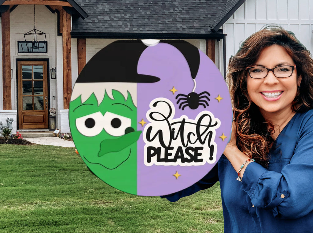 A smiling woman in glasses holds a round sign with a cartoon witch face, spider, and the words Witch Please! in front of a modern house with a lawn.