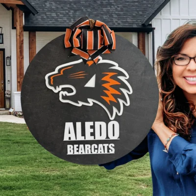 A smiling woman with long brown hair and glasses holds an Aledo Bearcat Mascot Door Hanger Door Sign, featuring the orange and black bearcat logo, while standing in front of a modern house with a green lawn.