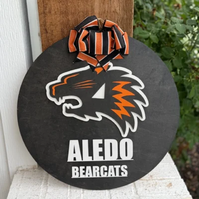 The Aledo Bearcat Mascot Door Hanger Door Sign is a round black sign with the orange and white Aledo Bearcat logo, Aledo Bearcats text, and an orange-black striped ribbon, perfect for displaying school spirit outdoors.