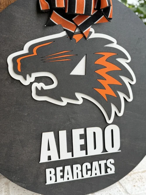The Aledo Bearcat Mascot Door Hanger Door Sign is a round black sign featuring a bold orange, white, and black bearcat logo with Aledo Bearcats below. Finished with a black and orange striped bow, it’s perfect for showcasing team spirit on your door.