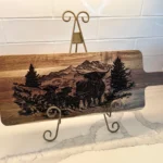 BBQ Grill Chart engraved Acacia Wood Cutting Board displayed on a decorative metal stand, set against a white tiled wall and marble countertop, featuring an intricate mountain, tree, and bison with calf scene.