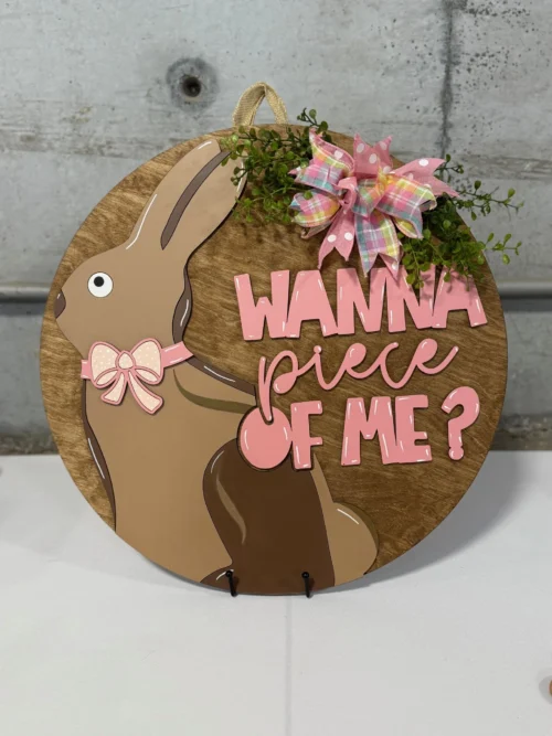 Round wooden sign with a brown bunny wearing a pink bow and a playful expression. Pink text reads Wanna piece of me? A pink and green plaid bow with greenery decorates the top right. Background is gray concrete.