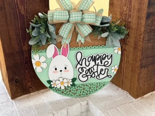 A round, pastel green Easter sign hangs on a wooden surface. It features a cartoon bunny with pink ears, surrounded by white flowers with yellow centers. The text Happy Easter is in black cursive. A burlap and green-checked ribbon bow is at the top, adorned with green leaves.