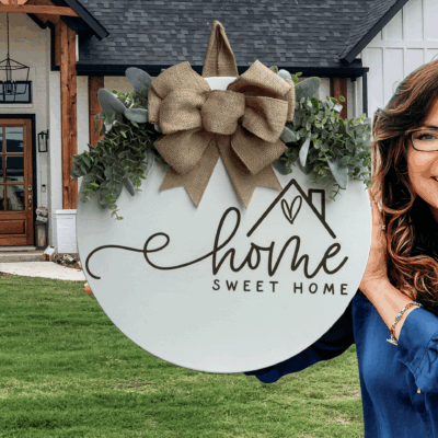 A smiling woman in glasses holds a decorative round sign with greenery and a bow that reads Home Sweet Home, standing in front of a modern house with a wooden front door and green lawn.
