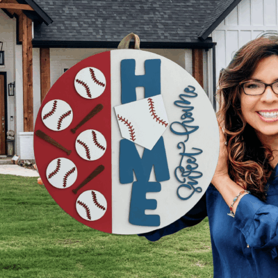 A smiling woman with long brown hair and glasses holds a round sign decorated with baseballs, bats, the word HOME, and Sweet Game, standing in front of a white house with a wooden porch.