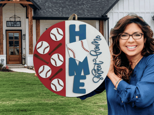 A smiling woman with long brown hair and glasses holds a round sign decorated with baseballs, bats, the word HOME, and Sweet Game, standing in front of a white house with a wooden porch.