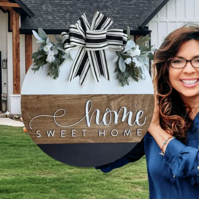 A smiling woman with wavy hair and glasses holds a round Home Sweet Home sign decorated with greenery and a striped bow, standing in front of a modern farmhouse-style home.