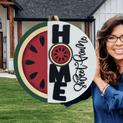 A smiling woman with long brown hair and glasses holds a round Home Sweet Home sign with a watermelon design in front of a modern farmhouse.