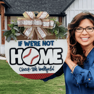 A smiling woman with long brown hair and glasses holds the If Were Not HOME Check the Ballfield Door Sign, Door Wreath, a round sign with a bow, greenery, and a baseball graphic highlighting HOME.