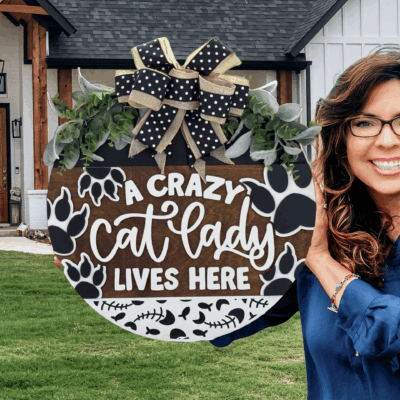 A smiling woman with wavy hair and glasses holds a round sign decorated with a bow, paw prints, and foliage that reads, A crazy cat lady lives here, standing outside a house with a wooden porch.