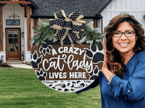 A smiling woman with wavy hair and glasses holds a round sign decorated with a bow, paw prints, and foliage that reads, A crazy cat lady lives here, standing outside a house with a wooden porch.