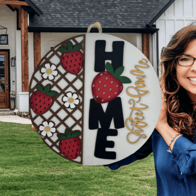A smiling woman with long brown hair and glasses holds a round Home Sweet Home sign decorated with strawberries and flowers, standing in front of a modern house with wood and white exterior.