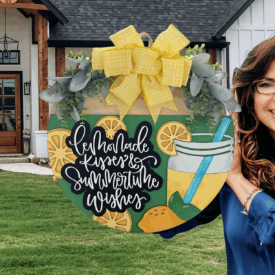 A smiling woman with long brown hair and glasses holds a large decorative sign reading “Lemonade Kisses & Summertime Wishes,” featuring lemons, a lemonade drink, greenery, and a yellow plaid bow, standing in front of a modern farmhouse.