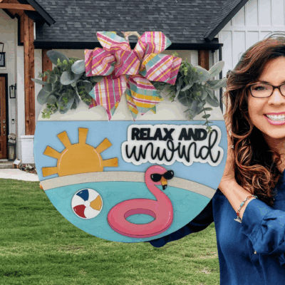 A smiling woman with long brown hair and glasses holds a round summer-themed sign that says Relax and Unwind, decorated with a sun, flamingo, beach ball, and a large colorful bow, standing in front of a house.