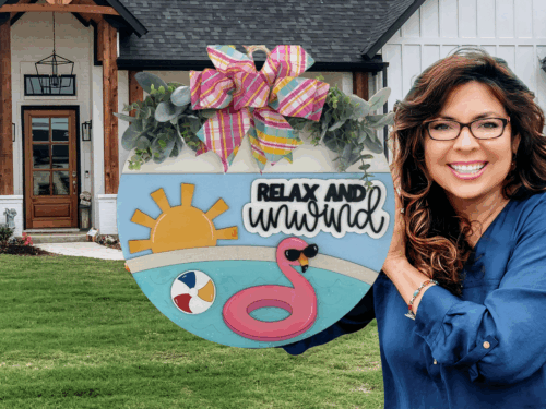A smiling woman with long brown hair and glasses holds a round summer-themed sign that says Relax and Unwind, decorated with a sun, flamingo, beach ball, and a large colorful bow, standing in front of a house.