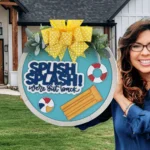 Woman smiling and holding a decorative sign. The sign is circular, blue, with SPLISH SPLASH! were out back in bold letters. It features a pool float, beach balls, and life rings. A yellow polka-dot ribbon and greenery adorn the top. The background shows a house with a wooden porch.