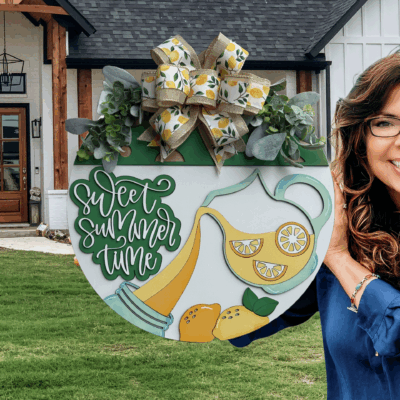 A smiling woman with wavy brown hair and glasses holds a round summer-themed sign with lemons, lemonade, and the words sweet summer time in front of a modern farmhouse.