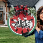 A woman with long hair and glasses stands outside a modern house holding a round sign. The sign resembles a watermelon slice with the text Sweet Summer written on it, adorned with a polka dot bow and greenery. The house has a wooden front door and white siding.