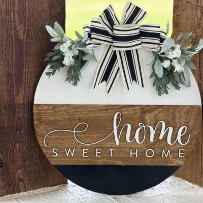 A round wooden sign with a striped black and white bow, greenery, and white flowers on top. The sign reads Home Sweet Home in white script across a brown wooden band.