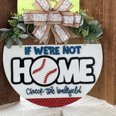 A round sign decorated with greenery and a bow featuring baseball designs reads, If were not home, check the ballfield. The letter O in home is styled as a baseball. The sign hangs on a wooden surface.