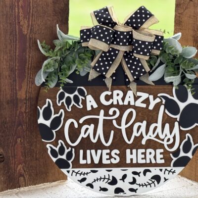 A round wooden sign with black paw prints, white bones, greenery, and a black polka-dot ribbon reads, A crazy cat lady lives here. The sign hangs on a rustic wooden background.