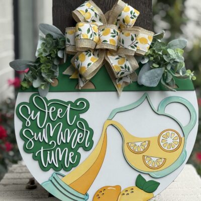 A decorative summer sign with a large bow, greenery, and lemons. The sign features a pitcher pouring lemonade and the words Sweet Summer Time in playful green lettering. Red flowers and a white brick wall are in the background.