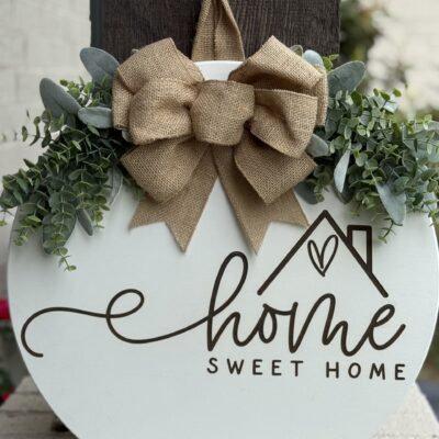 Round wooden sign with “Home Sweet Home” written in script, decorated with green leaves and a large burlap bow on top, hanging on a dark post with a white brick wall in the background.