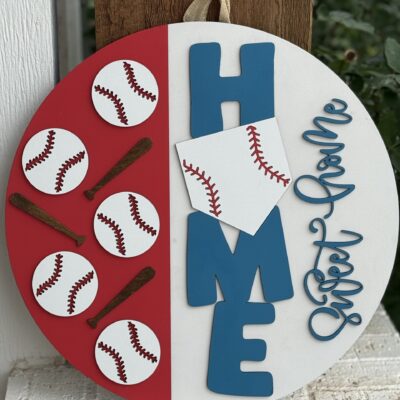 A round sign with baseballs and bats on a red half, and the word HOME in blue with a white home plate as the O. Sweet Home is written in blue script on the right side. The sign hangs by a loop at the top.