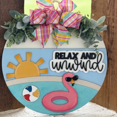 A round summer-themed door sign with faux greenery, a plaid bow, a sun, a pool with a pink flamingo float, a beach ball, and the words Relax and Unwind in black and white letters.