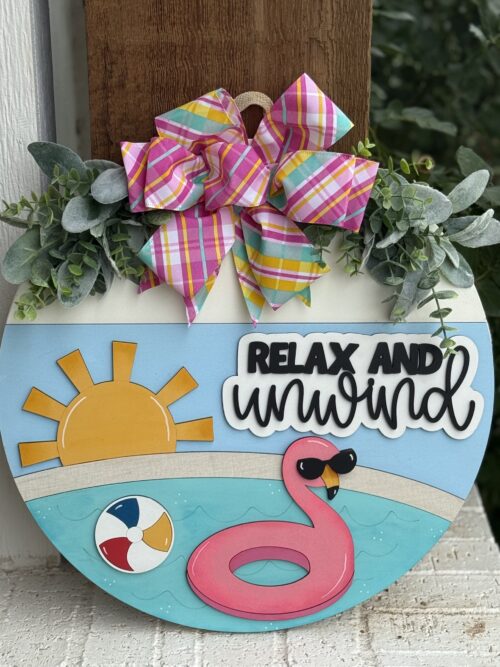 A round decorative sign features a pink inflatable flamingo, a beach ball, a bright sun, green leaves, and a colorful plaid bow. The sign reads RELAX AND unwind in bold and script fonts.
