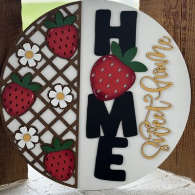 A round decorative sign features strawberries, white flowers, the word HOME in bold black letters with a strawberry for the O, and the phrase Sweet Home in cursive yellow on a white background.