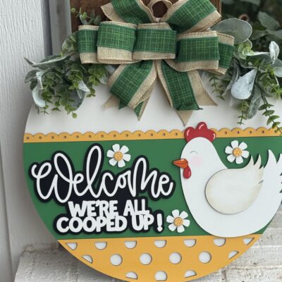 A round decorative sign with a cartoon chicken, flowers, and the text Welcome Were All Cooped Up! It features a green and yellow polka-dot pattern and is topped with a green and burlap bow and artificial greenery.