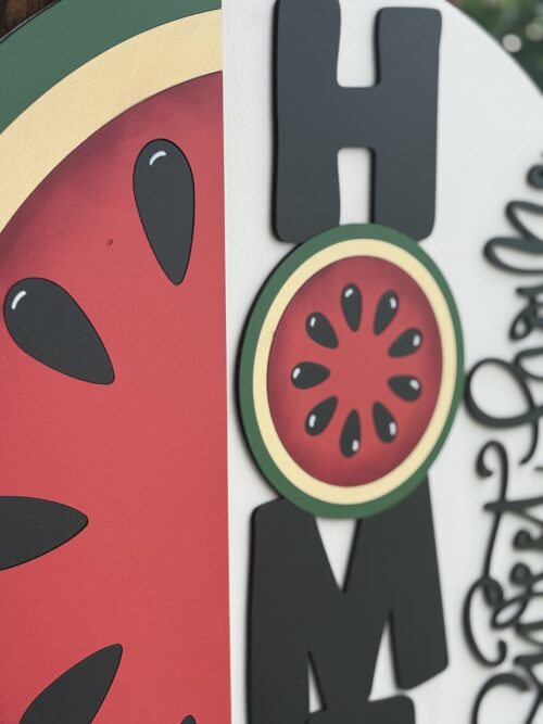A close-up of a decorative sign featuring the word HOME with a watermelon slice used as the letter O, and part of a handwritten Welcome phrase visible on the right side.