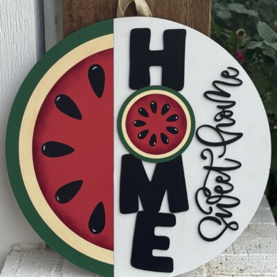 A round sign features a watermelon slice on the left and the word HOME vertically, with the O as a watermelon. Beside it, sweet home is written in cursive on a white background. Greenery is visible in the background.