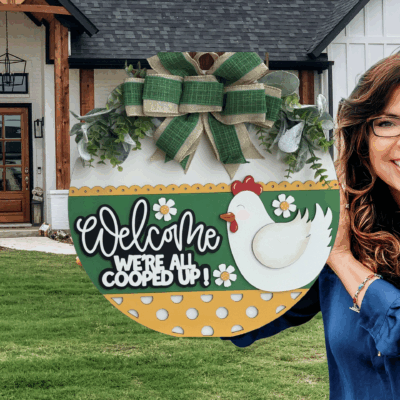 A smiling woman holds a round, decorative sign with a chicken, flowers, and the words Welcome, Were All Cooped Up! outside a modern farmhouse with a green lawn and wooden accents.