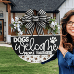 A smiling woman holds a round sign decorated with flowers and a striped bow that reads, Dogs Welcome, People Tolerated. She stands in front of a modern house with a green lawn.