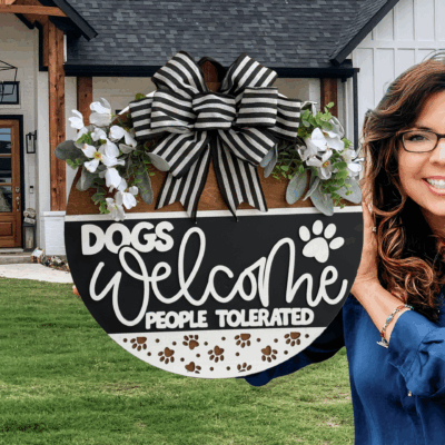 A smiling woman holds a round sign decorated with flowers and a striped bow that reads, Dogs Welcome, People Tolerated. She stands in front of a modern house with a green lawn.