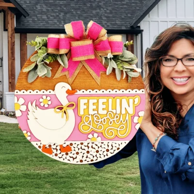 A smiling woman with glasses holds a decorative round sign featuring a duck, flowers, and the words Feelin Goosey, topped with a large pink and gold bow. A modern house is visible in the background.