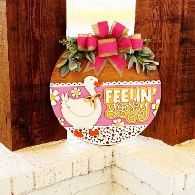 A round wooden sign with a white duck, pink and yellow flowers, and the text Feelin Lovely in yellow, decorated with greenery and a pink bow, hangs on a wooden post outside.