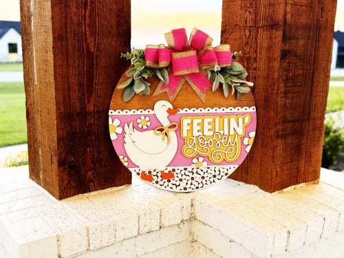 A round wooden sign with a white duck, pink and yellow flowers, and the text Feelin Lovely in yellow, decorated with greenery and a pink bow, hangs on a wooden post outside.