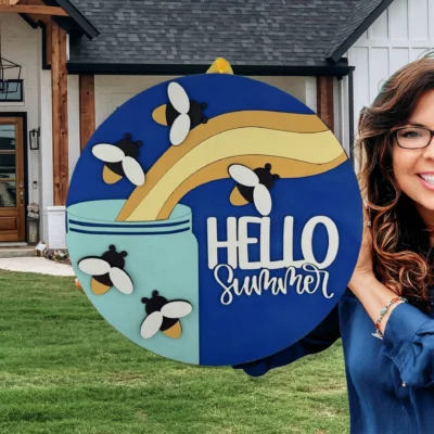 A smiling woman with glasses holds a colorful round sign that says HELLO summer with bees and honey art, standing outside a modern house with a manicured lawn.